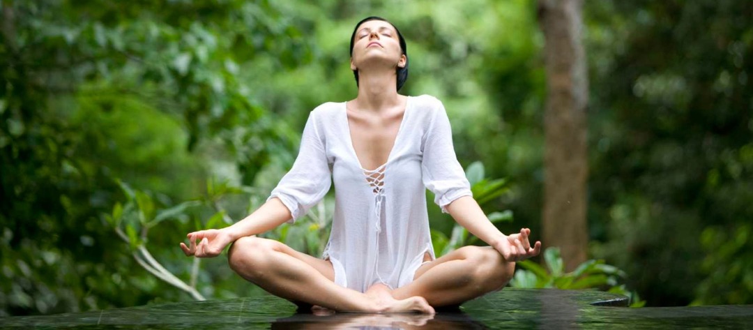 A woman sitting in a meditative pose looking up at the sky surrounded by nature