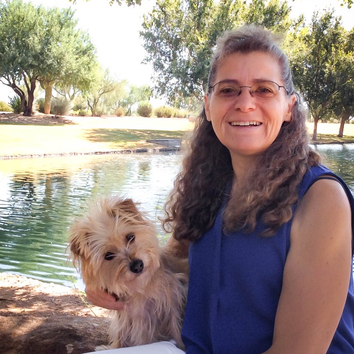 Dr. Evelyn outside with her dog on her lap sitting in front of a pond with trees on opposite bank.